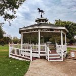 Union, OR 29 A white gazebo with a horse weathervane on top stands in a green park under a cloudy sky, surrounded by trees and grass. A ramp leads up to the gazebo’s wooden platform.