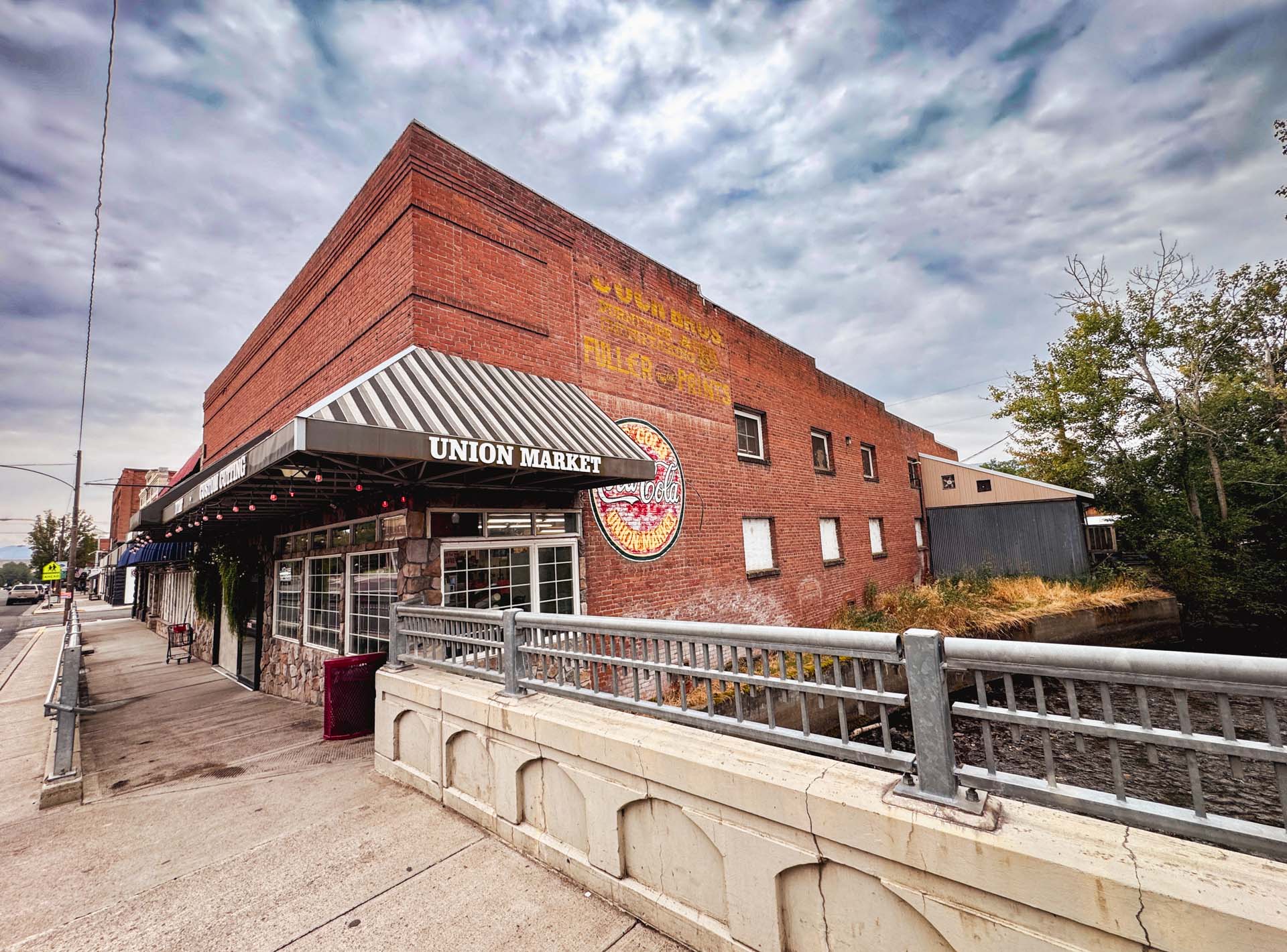 A brick building with a striped awning labeled Union Market stands beside a bridge. The sky is cloudy, and a faded mural is visible on the building’s upper wall. Trees and a small waterway run alongside.