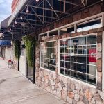 Union, OR 26 Sidewalk view of a stone-faced building with large windows and hanging plants; awning overhead, street and parked cars visible on the left.
