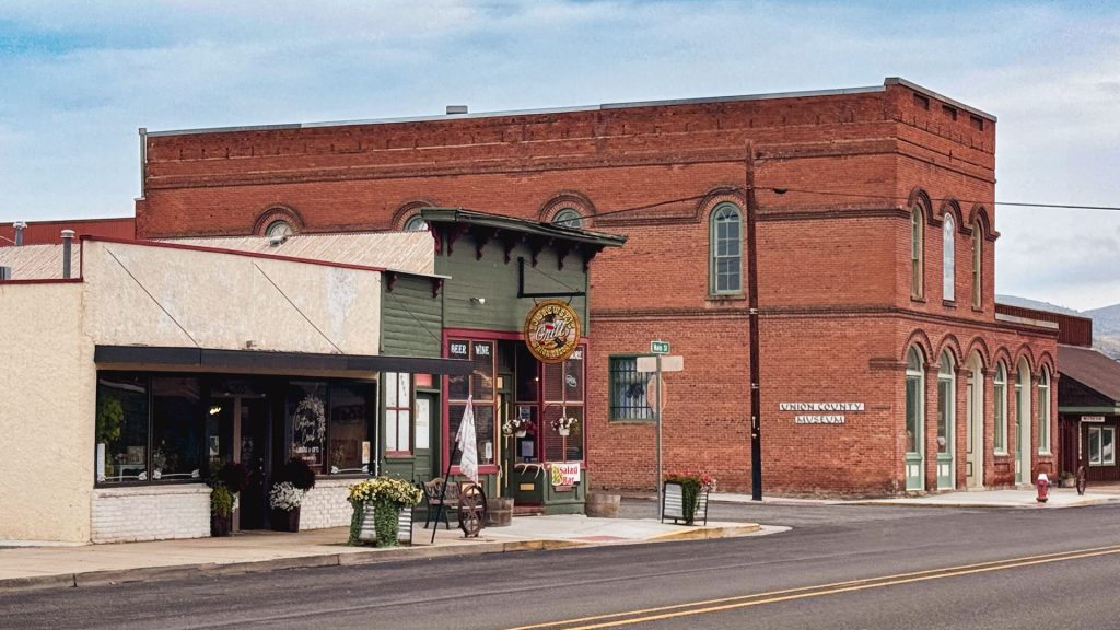 A quiet street corner in a small town with historic brick buildings, including a red brick structure labeled COUNTY PIONEER & HISTORICAL SOCIETY, and a green storefront with decorative elements and potted plants.