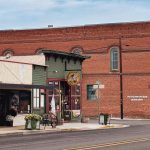 Union, OR 21 A quiet street corner in a small town with historic brick buildings, including a red brick structure labeled COUNTY PIONEER & HISTORICAL SOCIETY, and a green storefront with decorative elements and potted plants.
