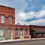 Union, OR 27 A small-town street features a historic red brick building with arched windows next to a one-story wooden structure, set under a cloudy sky with an American flag and greenery visible.