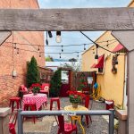 Union, OR 20 A cozy outdoor patio with red chairs, checkered tablecloths, potted plants, and string lights. The space is bordered by brick and yellow walls, with decorations and a hose visible in the background.