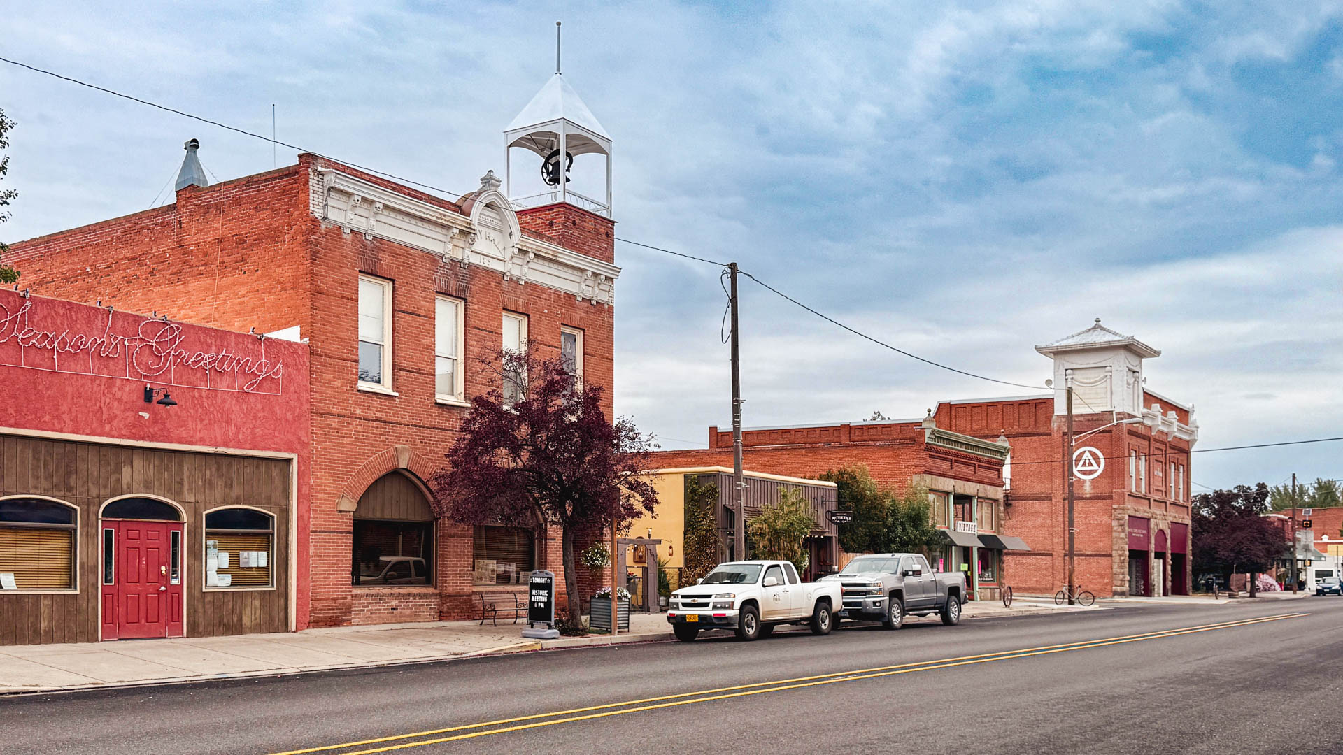 A small town main street with historic brick buildings, parked cars, and a cloudy sky. One building has a sign reading “Season’s Greetings” in cursive lights and another has a tall white clock tower.
