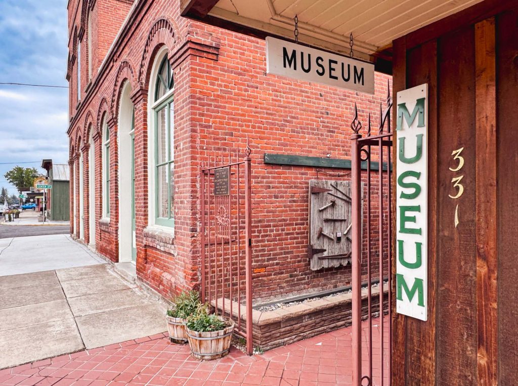 A red brick building on a street corner has two signs reading MUSEUM above the entrance and on the wall, with potted plants and large windows visible. The number 331 is displayed on the wooden wall.