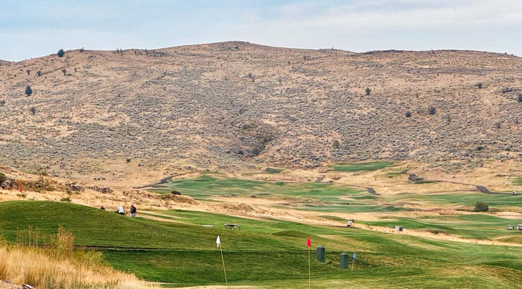 A wide view of a golf course with green fairways and flags, set against a backdrop of dry, rolling hills under a partly cloudy sky.