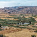 Union, OR 18 Aerial view of a sprawling golf course with green fairways, a pond, and scattered trees, surrounded by dry, brown hills and mountains under a cloudy sky. Several buildings sit near the edge of the course.