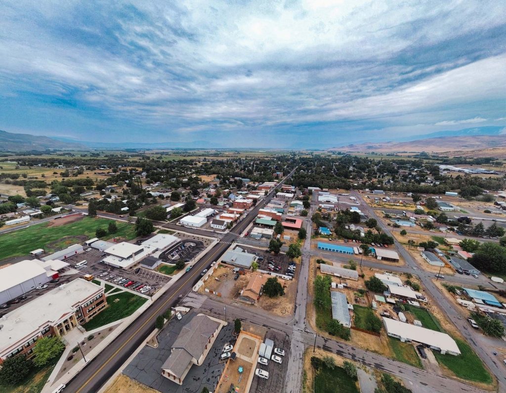 Aerial view of a small town with a main road running through the center, surrounded by houses, buildings, parking lots, and green spaces under a partly cloudy sky.