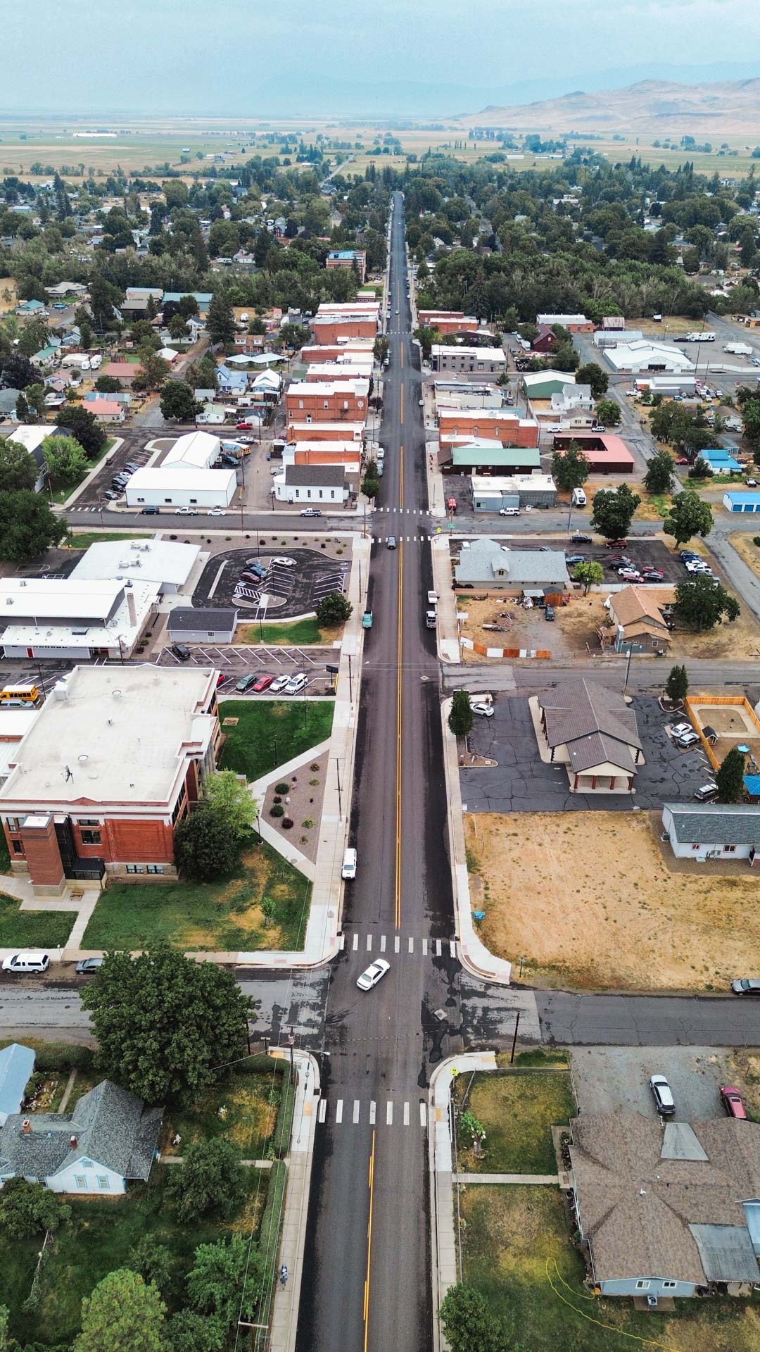 Aerial view of a small town with a straight road running through the center, lined with houses, buildings, trees, and parked cars on both sides. The landscape extends to distant hills under a hazy sky.