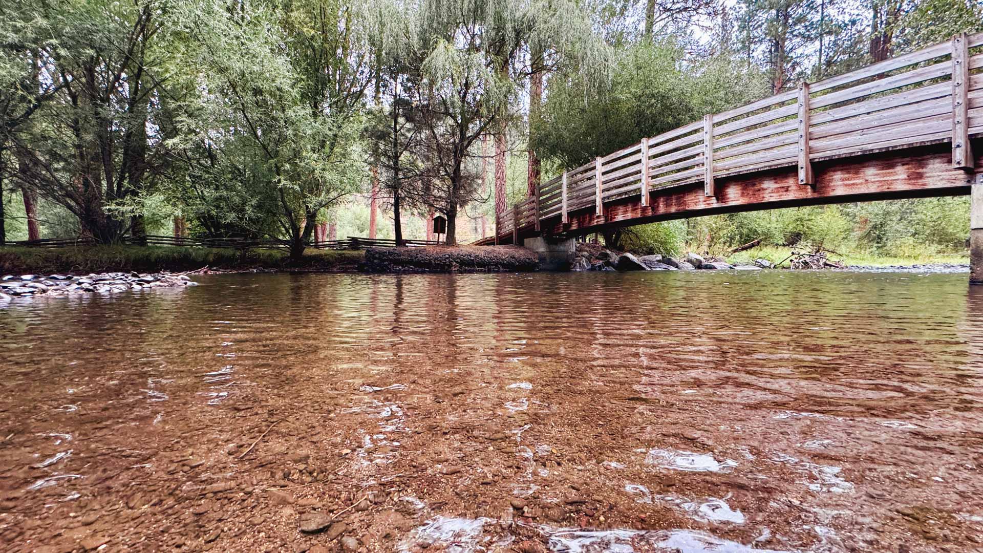 A wooden footbridge crosses over a shallow, clear creek surrounded by lush green trees and foliage, with rocks visible under the water and in the background.